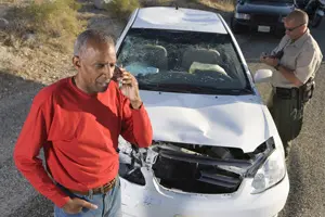a man talking on a cell phone in front of a damaged car